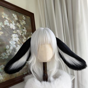 May include: A white wig featuring straight hair with bangs and two long, plush animal ears attached to the sides. The ears are black on the outer edges and white on the inner sections, creating a distinct contrast against the white hair.