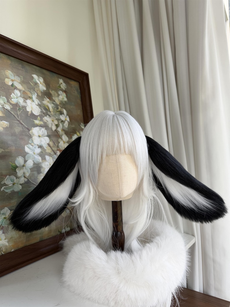 May include: A white wig featuring straight hair with bangs and two long, plush animal ears attached to the sides. The ears are black on the outer edges and white on the inner sections, creating a distinct contrast against the white hair.