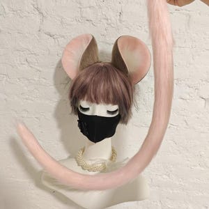May include: A mannequin head wearing a short brown wig, a black face mask, and a pearl necklace, featuring large brown and pink animal ears. A long, light pink faux fur tail is attached to the top of the head and curves downward.