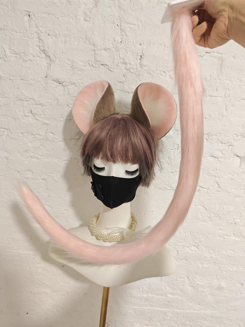 May include: A mannequin head wearing a short brown wig, a black face mask, and a pearl necklace, featuring large brown and pink animal ears. A long, light pink faux fur tail is attached to the top of the head and curves downward.