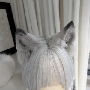 Puede incluir: Una peluca gris claro con orejas de lobo de piel a juego. La peluca tiene cabello liso, largo hasta los hombros con flequillo. Las orejas son grises con puntas negras. La peluca se exhibe en una cabeza de maniqu&iacute;.