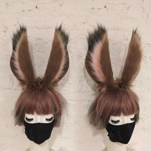 May include: Two mannequin heads wearing brown and pink faux fur animal ears with dark tips, paired with short pinkish-brown wigs featuring bangs. Each mannequin is wearing a solid black face mask with a small decorative detail on the side.