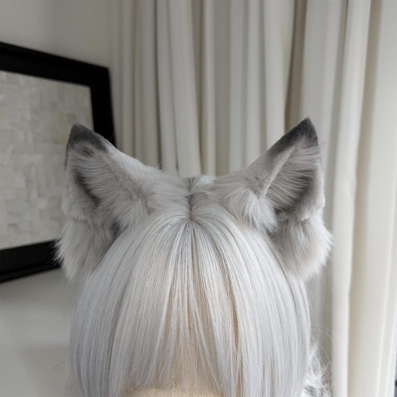 Puede incluir: Una diadema con orejas de animal de piel sint&eacute;tica gris claro y gris oscuro. Las orejas son erguidas y puntiagudas, con un color gris m&aacute;s oscuro en el interior y en las puntas. Las orejas se colocan sobre un fondo de pelo blanco.