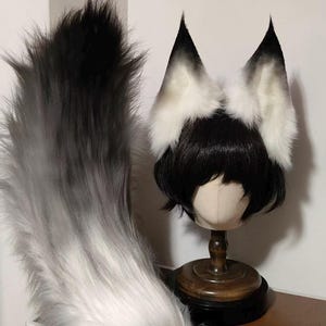 May include: A long, fluffy faux fur tail with a gradient from white at the base to black at the tip, paired with a set of tall, pointed animal ears in matching black and white fur, displayed on a black wig atop a wooden mannequin head stand.