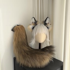 May include: A faux fur fox tail and ear set. The tail is a blend of brown and tan, with a black base. The ears are white, black, and tan. The set is displayed on a black surface.