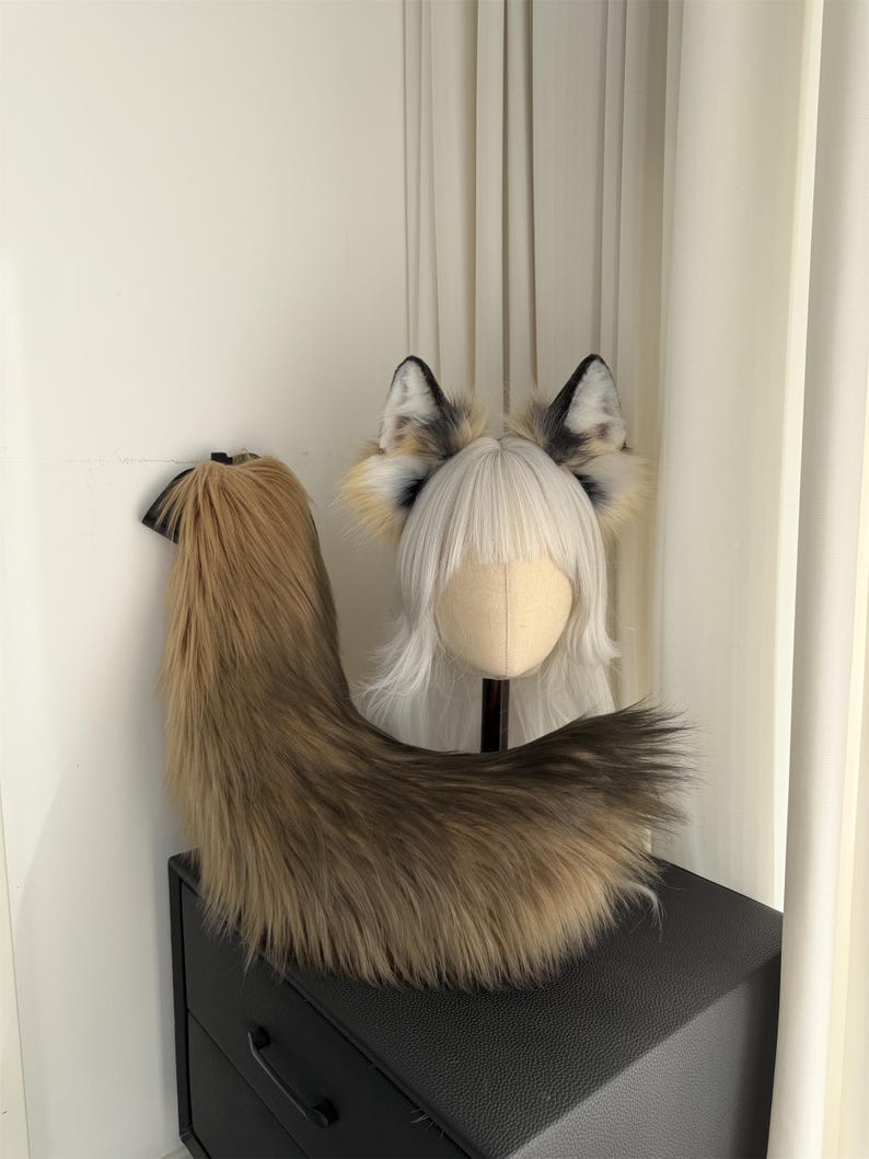 May include: A faux fur fox tail and ear set. The tail is a blend of brown and tan, with a black base. The ears are white, black, and tan. The set is displayed on a black surface.