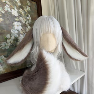 May include: A white wig with bangs styled on a mannequin head, featuring two long, plush, brown and white rabbit-style ears attached to the sides. A matching, fluffy, brown and white faux fur tail is positioned in front of the wig base.
