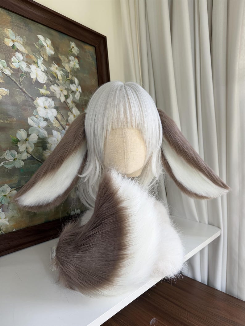 May include: A white wig with bangs styled on a mannequin head, featuring two long, plush, brown and white rabbit-style ears attached to the sides. A matching, fluffy, brown and white faux fur tail is positioned in front of the wig base.