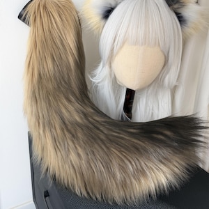 May include: A faux fur tail in shades of brown and black, with a black attachment. A wig head with a white wig and matching fox ears with black, white, and yellow accents is also visible. The items are likely part of a cosplay or costume set.