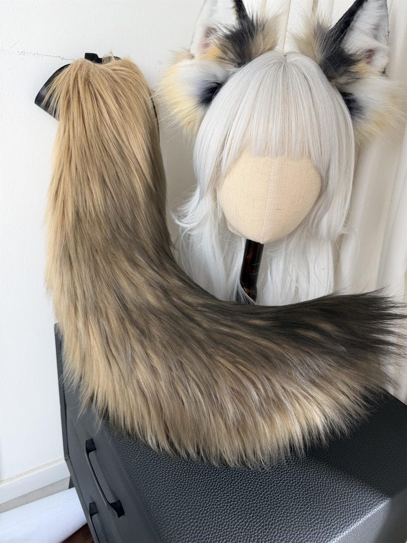 May include: A faux fur tail in shades of brown and black, with a black attachment. A wig head with a white wig and matching fox ears with black, white, and yellow accents is also visible. The items are likely part of a cosplay or costume set.