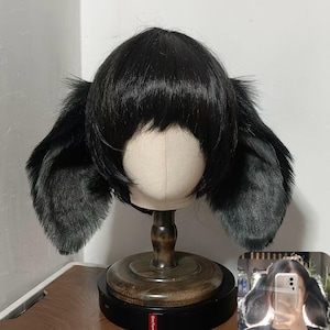 May include: A black wig with straight bangs and long, soft, furry animal-like ears attached to the sides. The wig is displayed on a beige mannequin head mounted on a dark wooden stand, featuring a smooth texture and a layered, rounded cut.