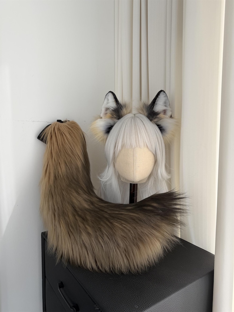 May include: A faux fur fox tail and ear set. The tail is brown and tan, with a black ribbon attachment. The ears are white, black, and tan, attached to a wig head with white hair. The set is designed for cosplay or costume use.