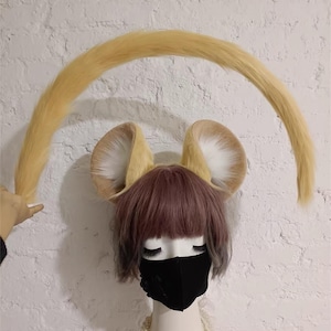 May include: A mannequin head wearing a short mauve wig, a black face mask, and a white beaded necklace. Attached to the head are two large, upright, tan and white faux fur animal ears and a long, curved, tan faux fur tail held in an arch.