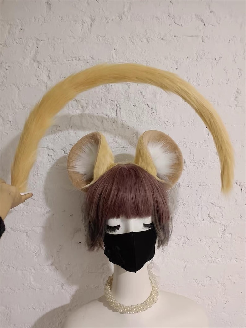 May include: A mannequin head wearing a short mauve wig, a black face mask, and a white beaded necklace. Attached to the head are two large, upright, tan and white faux fur animal ears and a long, curved, tan faux fur tail held in an arch.
