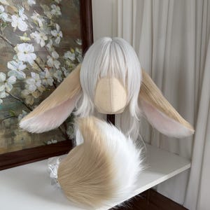 May include: A white wig with straight bangs and long side layers, featuring two large, pointed, tan and white faux fur animal ears attached to the sides. A matching, fluffy, tan and white faux fur tail is positioned in front of the wig.