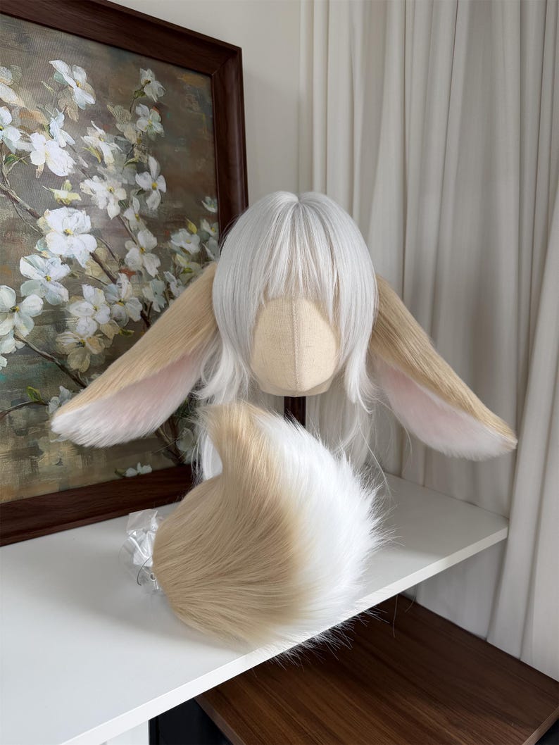 May include: A white wig with straight bangs and long side layers, featuring two large, pointed, tan and white faux fur animal ears attached to the sides. A matching, fluffy, tan and white faux fur tail is positioned in front of the wig.