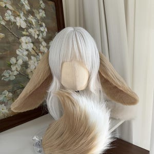 May include: A white wig with straight bangs and long layers, featuring two large, plush, tan-colored animal ears attached to the sides. A matching, fluffy, tan and white faux fur tail is positioned in front of the wig on a mannequin head.