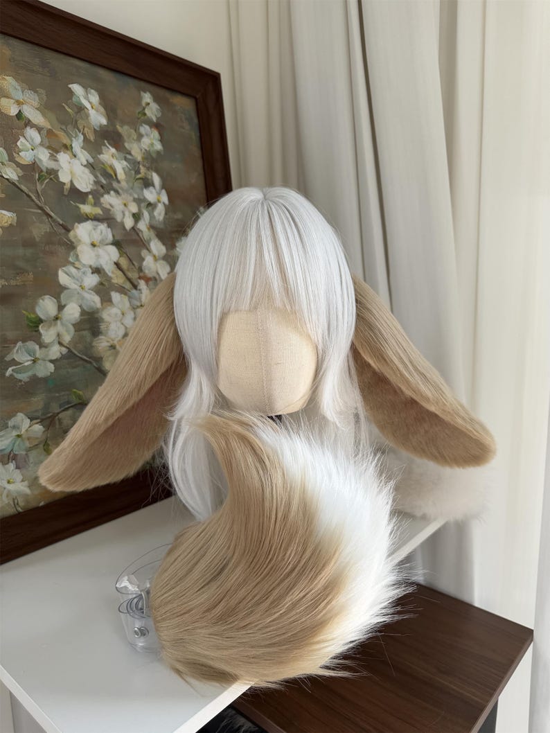 May include: A white wig with straight bangs and long layers, featuring two large, plush, tan-colored animal ears attached to the sides. A matching, fluffy, tan and white faux fur tail is positioned in front of the wig on a mannequin head.