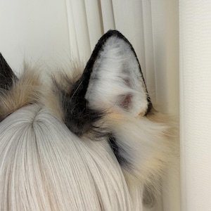 May include: A pair of faux fur wolf ears in black, white, and tan. The ears are attached to a headband and are designed to be worn on the head. The fur is fluffy and textured, with a realistic appearance. The ears are a cosplay or costume accessory.