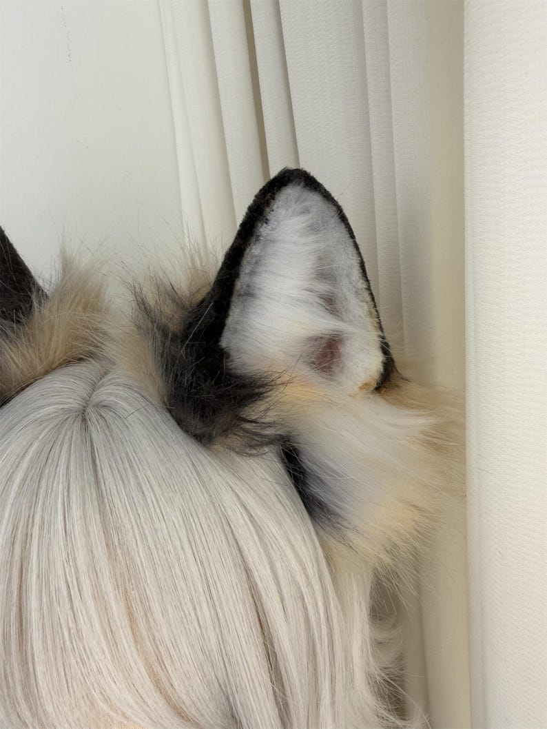 May include: A pair of faux fur wolf ears in black, white, and tan. The ears are attached to a headband and are designed to be worn on the head. The fur is fluffy and textured, with a realistic appearance. The ears are a cosplay or costume accessory.
