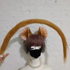 May include: A mannequin head wearing a short, layered, dusty-pink wig, a black face mask, and a white pearl necklace. Attached to the wig are two large, rounded, tan and pink animal ears and a long, curved, golden-brown tail arching overhead.