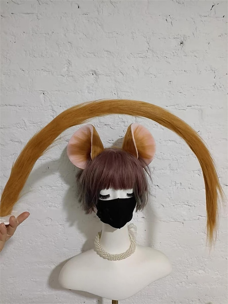 May include: A mannequin head wearing a short, layered, dusty-pink wig, a black face mask, and a white pearl necklace. Attached to the wig are two large, rounded, tan and pink animal ears and a long, curved, golden-brown tail arching overhead.