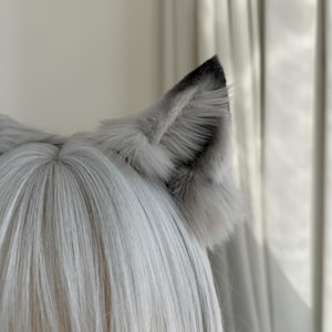Puede incluir: Un par de orejas de lobo de piel sint&eacute;tica gris unidas a una peluca. Las orejas son de color gris claro con puntas grises m&aacute;s oscuras y una textura esponjosa. La peluca tiene pelo largo, liso y gris claro.