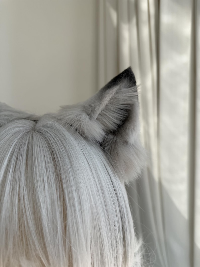 Puede incluir: Un par de orejas de lobo de piel sint&eacute;tica gris unidas a una peluca. Las orejas son de color gris claro con puntas grises m&aacute;s oscuras y una textura esponjosa. La peluca tiene pelo largo, liso y gris claro.