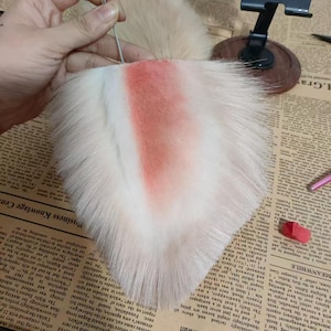 May include: A triangular faux fur ear accessory featuring long, cream-colored synthetic fibers with a soft, airbrushed coral-pink gradient concentrated in the center. A thin, silver-toned metal wire pin is inserted into the top edge of the base.
