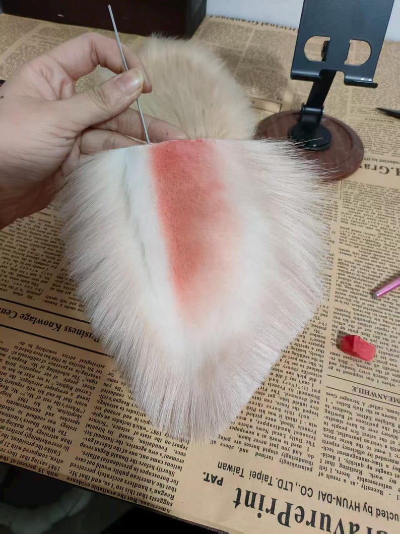 May include: A triangular faux fur ear accessory featuring long, cream-colored synthetic fibers with a soft, airbrushed coral-pink gradient concentrated in the center. A thin, silver-toned metal wire pin is inserted into the top edge of the base.