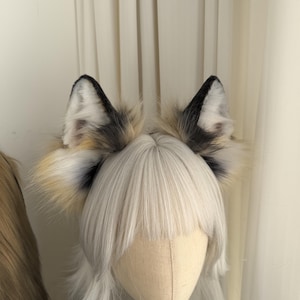 May include: A pair of faux fur fox ears with black tips and a mix of white, tan, and grey fur. The ears are attached to a headband and are displayed on a mannequin head with a white wig.