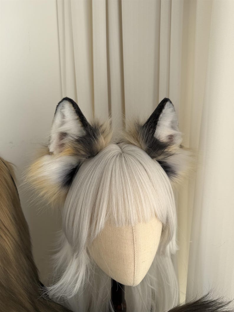May include: A pair of faux fur fox ears with black tips and a mix of white, tan, and grey fur. The ears are attached to a headband and are displayed on a mannequin head with a white wig.