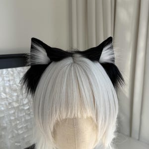 Op de afbeelding: Zwart-witte kattenoren-hoofdband. De oren zijn zwart met wit, pluizig haar aan de binnenkant. De hoofdband wordt gedragen op een mannequinhoofd met wit haar.