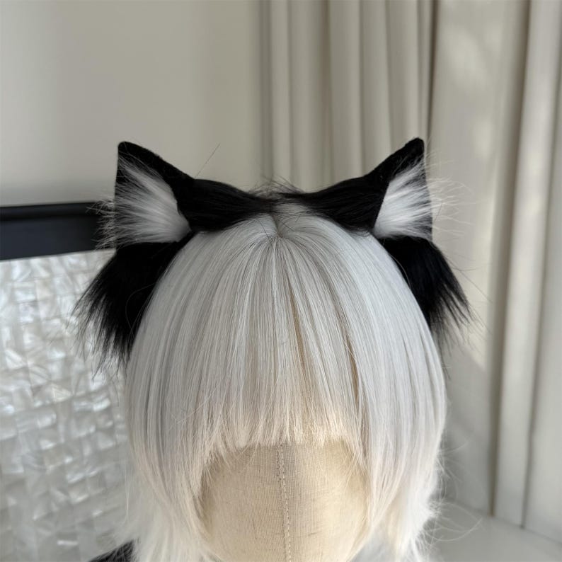 Op de afbeelding: Zwart-witte kattenoren-hoofdband. De oren zijn zwart met wit, pluizig haar aan de binnenkant. De hoofdband wordt gedragen op een mannequinhoofd met wit haar.