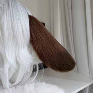 May include: A pair of animal-inspired ears featuring long, straight white synthetic hair and a single prominent, smooth brown ear flap made of faux fur. The design is sleek, with the white hair layered and the brown ear extending outward.