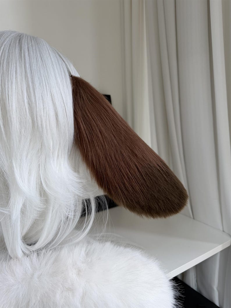 May include: A pair of animal-inspired ears featuring long, straight white synthetic hair and a single prominent, smooth brown ear flap made of faux fur. The design is sleek, with the white hair layered and the brown ear extending outward.