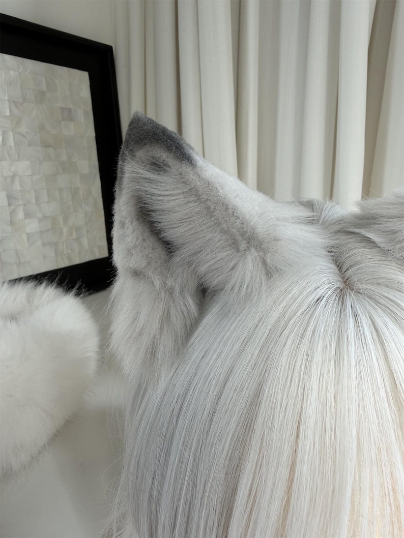 Puede incluir: Primer plano de un par de orejas de lobo de piel sint&eacute;tica gris y blanca unidas a una peluca con pelo largo, liso y gris claro. Las orejas tienen un interior gris m&aacute;s oscuro y son esponjosas. Un accesorio blanco y esponjoso es visible a la izquierda.