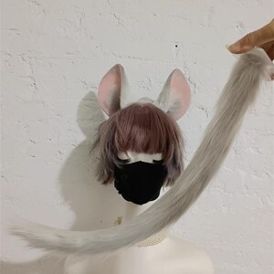 May include: A set of light gray faux fur animal ears and a matching long, slender faux fur tail. The ears feature soft pink inner detailing and are positioned atop a mannequin head wearing a short, layered brown wig and a solid black face mask.