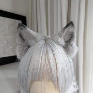 Puede incluir: Una diadema con orejas de gato de piel sint&eacute;tica gris y blanca. Las orejas son esponjosas con puntas grises m&aacute;s oscuras. Las orejas est&aacute;n colocadas sobre una peluca con pelo largo, liso y plateado.