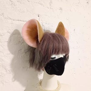 May include: A mannequin head wearing a black face mask with subtle embossed text, topped with large, upright, plush animal ears. The ears feature a soft, light pink inner surface and a textured, golden-brown outer surface, set against a short, layered, light purple wig.