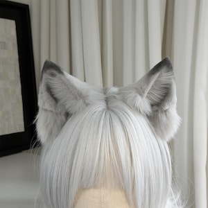 Puede incluir: Un par de orejas de lobo de piel sint&eacute;tica gris unidas a una diadema. Las orejas tienen un interior gris m&aacute;s oscuro y se colocan sobre un fondo de pelo blanco. Las orejas son esponjosas y detalladas.