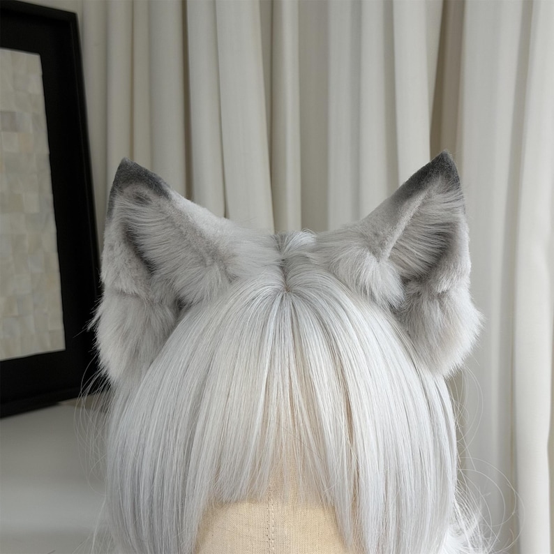 Puede incluir: Un par de orejas de lobo de piel sint&eacute;tica gris unidas a una diadema. Las orejas tienen un interior gris m&aacute;s oscuro y se colocan sobre un fondo de pelo blanco. Las orejas son esponjosas y detalladas.