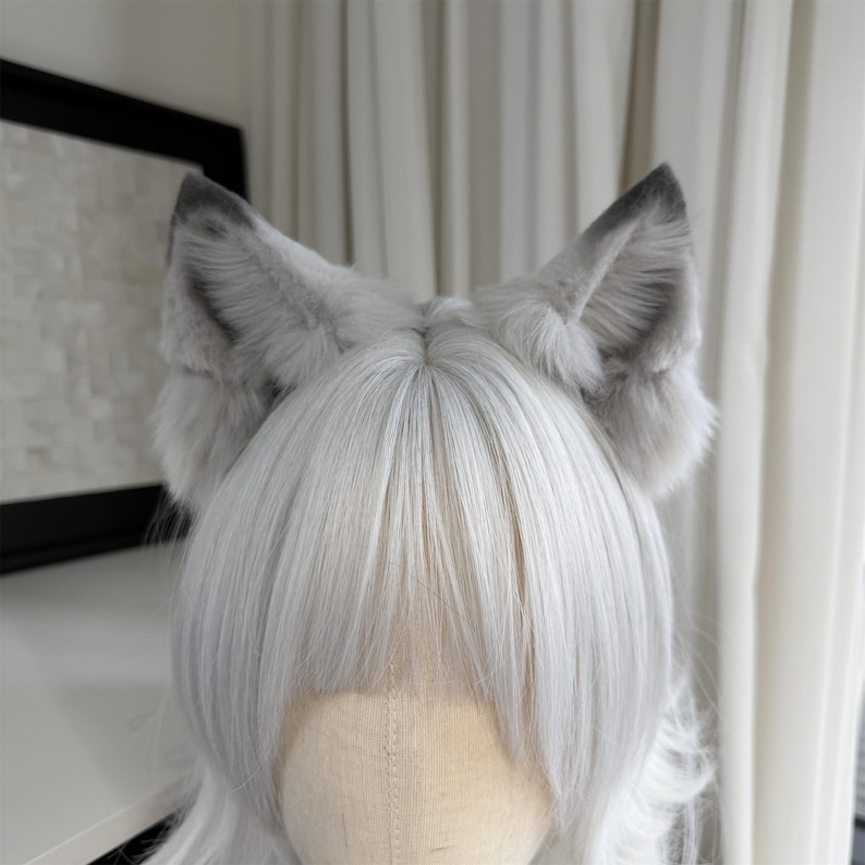 Puede incluir: Un par de orejas de lobo de piel sint&eacute;tica gris y blanca unidas a una diadema. Las orejas tienen una textura esponjosa y est&aacute;n rematadas con gris m&aacute;s oscuro. Las orejas est&aacute;n colocadas sobre una cabeza de maniqu&iacute; con pelo blanco.