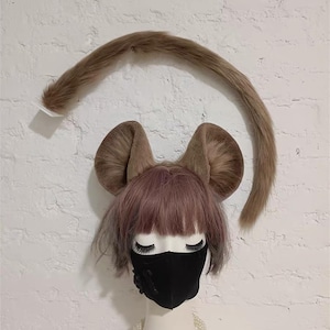 May include: A mannequin head wearing a light brown faux fur mouse ear headband and a matching long faux fur tail. The mannequin has short, light purple hair, a black face mask with a small black flower detail, and a white faux pearl necklace.