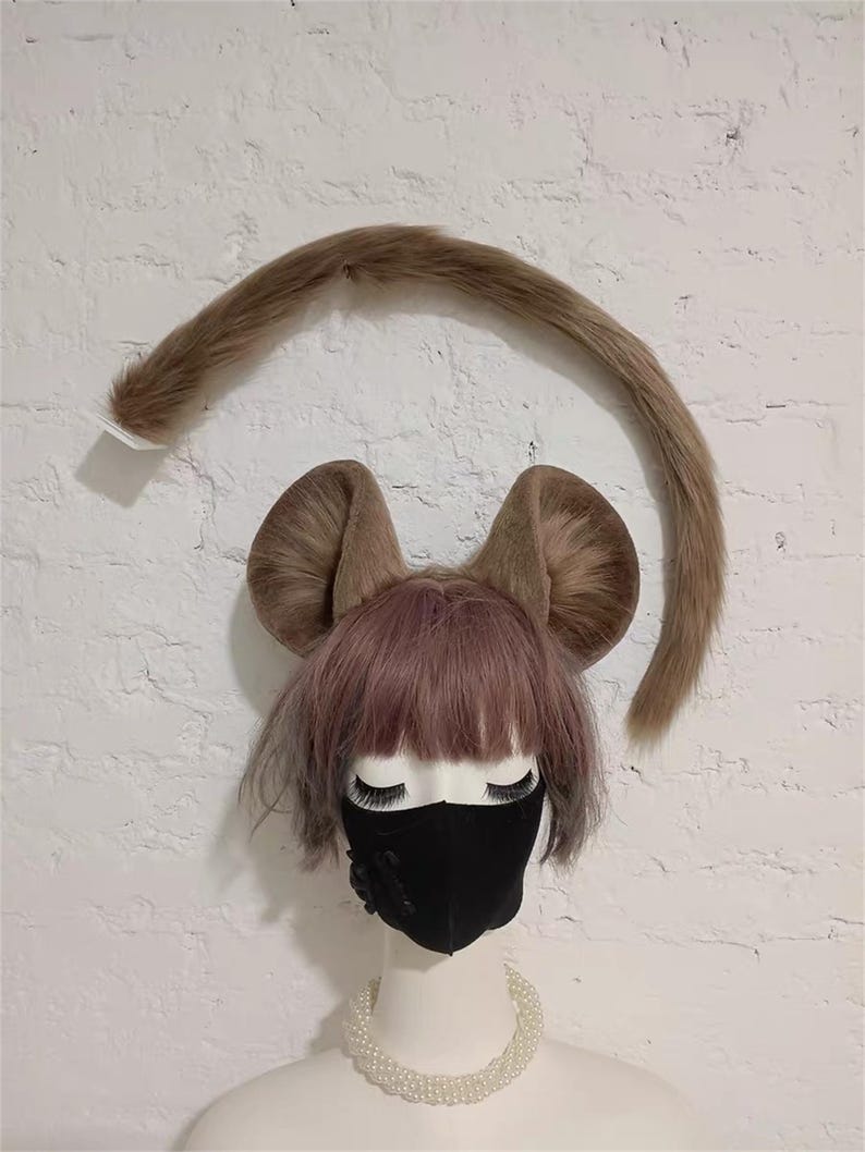May include: A mannequin head wearing a light brown faux fur mouse ear headband and a matching long faux fur tail. The mannequin has short, light purple hair, a black face mask with a small black flower detail, and a white faux pearl necklace.