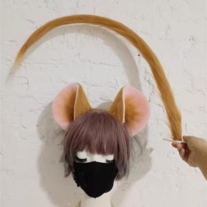 May include: A set of faux fur animal ears and a long, curved tail in a golden-brown hue. The ears feature soft pink inner detailing and are attached to a wig with short, layered, light brown hair. A solid black face mask covers the lower face.