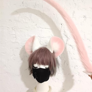 May include: A set of plush mouse ears in white and soft pink, paired with a long, matching pink faux fur tail. The ears are attached to a headband worn on a mannequin head with short brown hair, a black face mask, and a white pearl necklace.