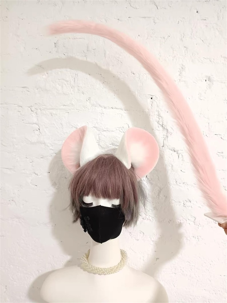 May include: A set of plush mouse ears in white and soft pink, paired with a long, matching pink faux fur tail. The ears are attached to a headband worn on a mannequin head with short brown hair, a black face mask, and a white pearl necklace.