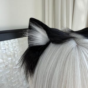 Op de afbeelding: Zwarte en witte kattenoren van imitatiebont. De oren zijn aan de buitenkant zwart en aan de binnenkant wit. De oren zijn bevestigd aan een zwarte hoofdband. De pruik heeft lang, steil, wit haar.