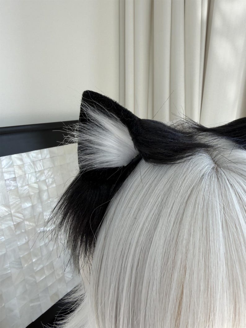 Op de afbeelding: Zwarte en witte kattenoren van imitatiebont. De oren zijn aan de buitenkant zwart en aan de binnenkant wit. De oren zijn bevestigd aan een zwarte hoofdband. De pruik heeft lang, steil, wit haar.